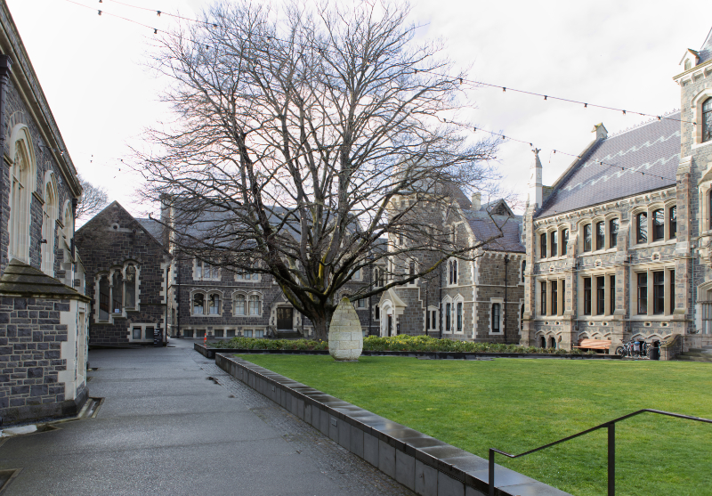 A grassed raised area with a large leafless tree and egg shaped sculpture is surrounded by  a footpath and gothic revival stone buildings in excellent condition