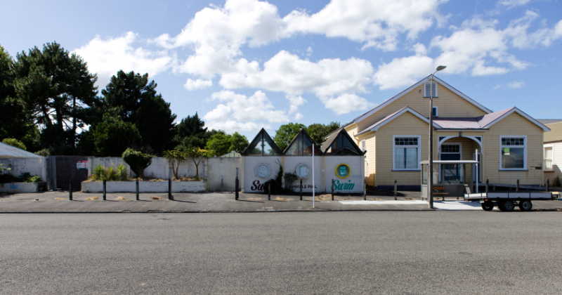 a sunny day photo of the street frontage of GCUR. Left the former Gonville swimming pool stucco wall with peaked rooves and porthole windows. Right a waetherboard clad community hall frontage