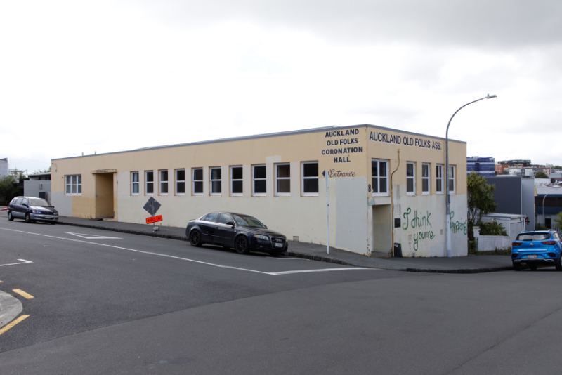 An off white and pale yellow painted one-story flat-roofed 1950s building with a painted name: Auckland Old Folks Coronation Hall. The building has been tagged with gree graffiti saying I think you're amazing