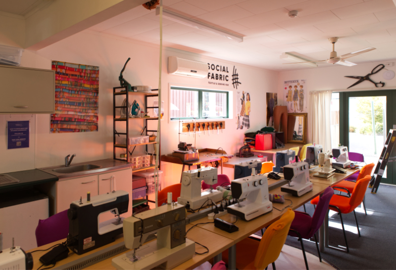 A interior photo showing a long table with nine sewing machines and chairs. The words Social Fabric are painted on the wall.